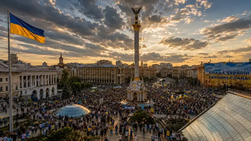 Kyiv’s Independence Square at sunset, filled with a large crowd around the Independence Monument, with a Ukrainian flag flying prominently in the foreground.