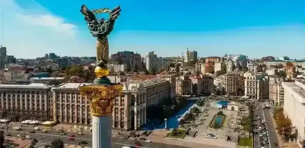 Kyiv’s Independence Square at sunset, filled with a large crowd around the Independence Monument, with a Ukrainian flag flying prominently in the foreground.